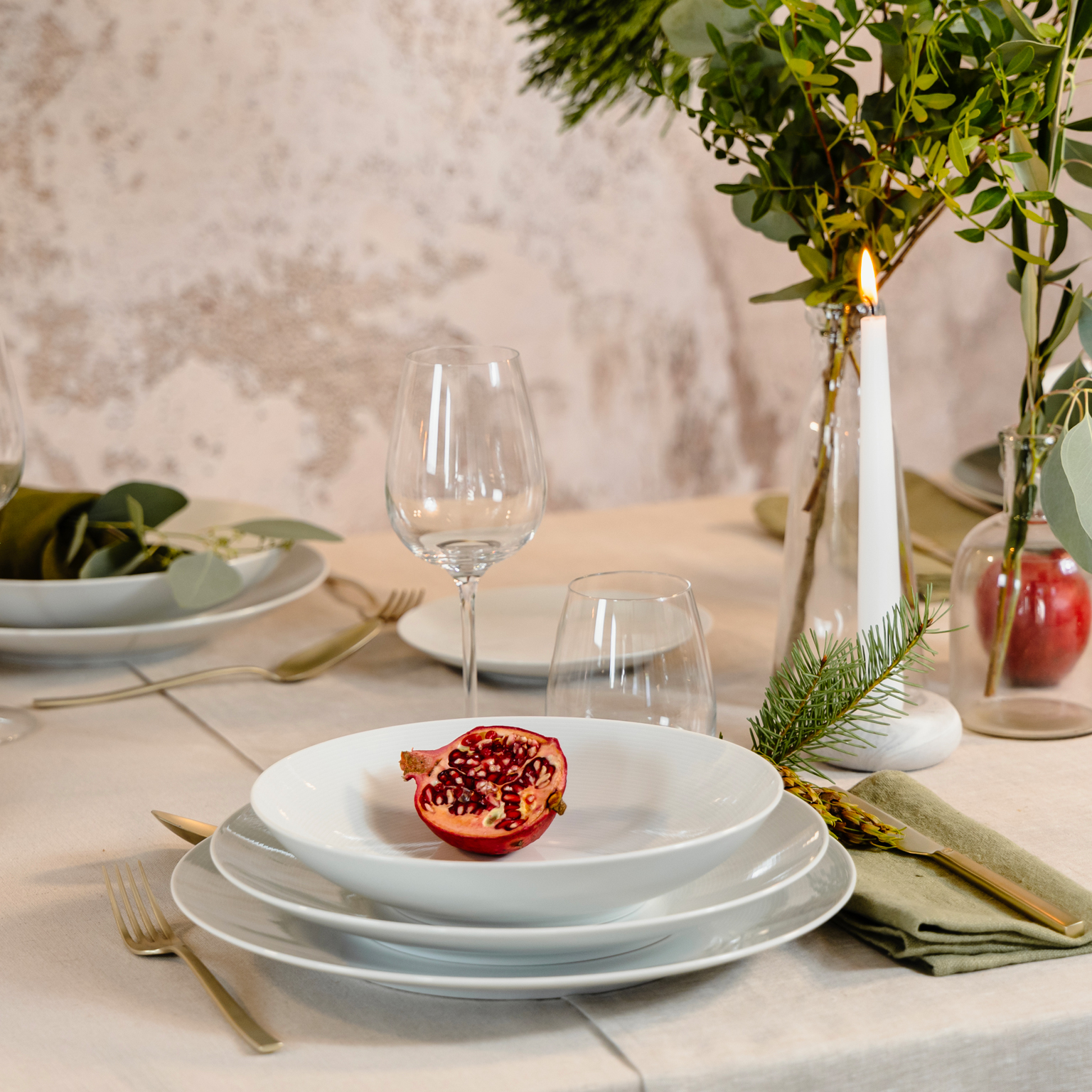 Thomas Loft White soup plate, dinner plate and charger plate stacked on top of each other with half a pomegranate as a decoration and wine glass, candle and bouquet of green branches in the background